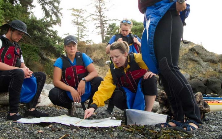 a group of people wearing life jackets examine a map that is spread out on a rocky shore.